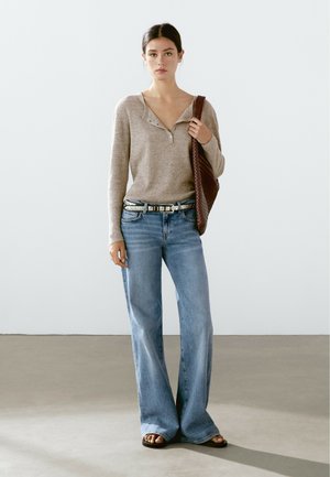 Braun gerippte Langarmbluse, hellblaue weit geschnittene Jeans, silberne Akzentgürtel und braune geflochtene Handtasche. Sandalen mit breiten Riemen und freiliegenden Zehen.