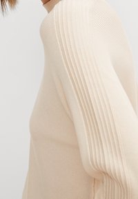 Maglione beige a coste con design aderente e maniche lunghe, caratterizzato da dettagli di tessuto testurizzato lungo le maniche e il collo.