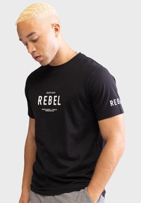 Crna pamučna majica s kratkim rukavima, s bijelim natpisima "REDEFINIRANO" i "REBEL" na prednjoj strani i rukavu, glatke teksture.