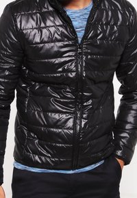 Black puffer jacket, quilted design with shiny texture, full zipper, standing collar, worn over a blue striped shirt, with side pockets.