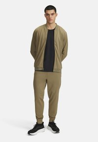 Sports tracksuit i olivengrøn, der har en lynlås bomberjakke og tætsiddende joggers, parret med sorte sneakers. Glat stoftekstur.