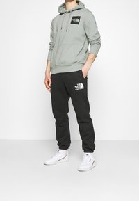 Sudadera gris clara con un bolsillo frontal y logotipo, combinada con pantalones de chándal negros que tienen un logotipo blanco, y zapatillas blancas.