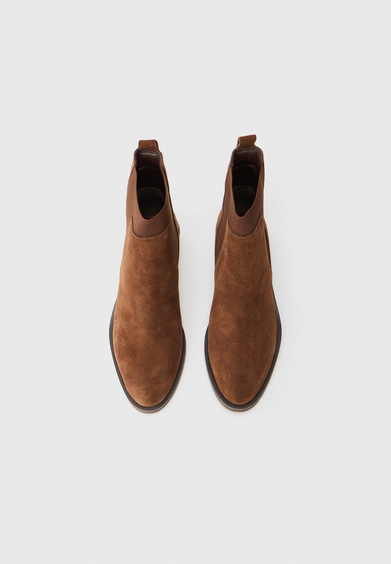 Braune Wildlederstiefeletten mit einem glatten Lederkragen, die über geschwungene Öffnungen und ein abgerundetes Zehendesign auf einer flachen Sohle verfügen.