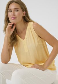 Top sin mangas amarillo con escote en V, hecho de una tela ligera y ligeramente texturizada. Presenta sutiles pliegues cerca de los hombros. Combínalo con pantalones blancos.