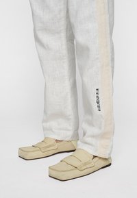 Lichte linnen broek met een gestructureerde witte afwerking, voorzien van een beige zijstreep en bedrukte cijfers. Beige instapschoenen met vierkante neuzen.