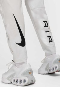 Hvide Nike bukser med et sort swoosh-design og lynlåsdetalje, parret med hvide sneakers med synlig air-dæmpning og logo.