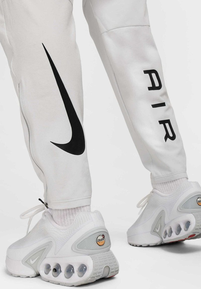 Pantalon Nike blanc avec un design de swoosh noir et des détails de fermeture éclair, associé à des baskets blanches avec un amorti visible et un logo.