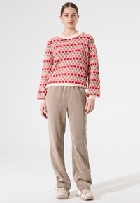 Femme debout portant un pull à manches longues à motifs rouge et beige, un pantalon en velours côtelé beige et des baskets blanches épaisses sur un fond blanc.