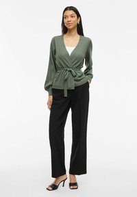 Cárdigan verde con mangas largas abullonadas, cintura anudada y puños acanalados, combinado con pantalones de pierna ancha negros y sandalias de tacón negras.