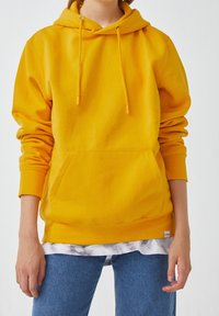 Personne portant un sweat à capuche jaune moutarde avec poche avant et manches retroussées, sur une chemise blanche à motifs et un jean bleu, debout les mains détendues.