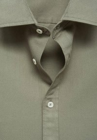 Camicetta di un verde oliva leggero con una colletto a bottoni. Presenta due bottoni bianchi all'altezza del collo e un design strutturato.