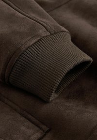 Braunes Wildledergewebe mit geripptem Strickbündchen. Glatte Textur, saubere Nähte, die eine Nahaufnahme des Ärmeldesigns und der Details präsentieren.