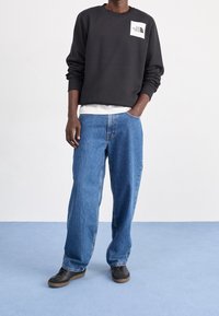 Sudadera negra con logo en el pecho, camiseta gris clara debajo, jeans de denim azul holgados y zapatillas negras con suela de goma.