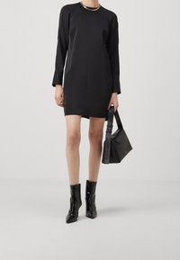 Frau in einem schwarzen langen Minikleid, schwarzen lackierten Stiefeletten, mit einer goldenen Halskette und einer schwarzen Handtasche von Calvin Klein.