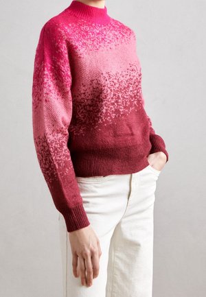 Personne portant un pull en tricot dégradé du rose au bordeaux avec un petit motif tacheté, associé à un pantalon blanc, main dans la poche.