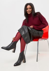Schwarze Stiefeletten mit spitzer Zehe und gemustertem Blockabsatz, kombiniert mit einem schwarzen Lederrock und einem kastanienbraunen Pullover.