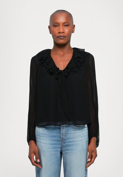 Blouse noire avec un col volanté et des liens au niveau du décolleté. Manches longues en dentelle avec une coupe ample. Associée à un jean clair.