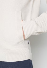 Main dans la poche latérale d'un pull blanc à manches longues côtelé avec une étiquette noire « BOSS », associé à un pantalon bleu foncé.