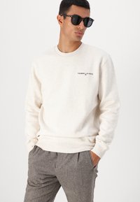 Svetlo béžová mikina s okrúhlym výstrihom a rebrovanými manžetami, na hrudi s malým čiernym a červeným logom "TOMMY JEANS".