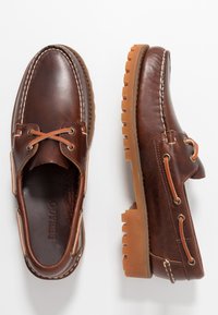 Braune Lederschuhe für Boote mit weißer Stickerei, runder Spitze und Gummisohle mit Zickzackmuster. Die Schnürsenkel sind hellbraun und durch metallische Ösen gefädelt.