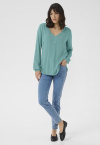 Light teal long-sleeve blouse with a V-neck and side slits, paired with blue skinny jeans and black flat shoes. Smooth texture throughout.