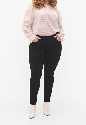 Kobieta ubrana w jasnoróżową, teksturowaną bluzkę z długim rękawem, czarne dopasowane spodnie i beżowe botki, stojąca z rękami w kieszeniach.