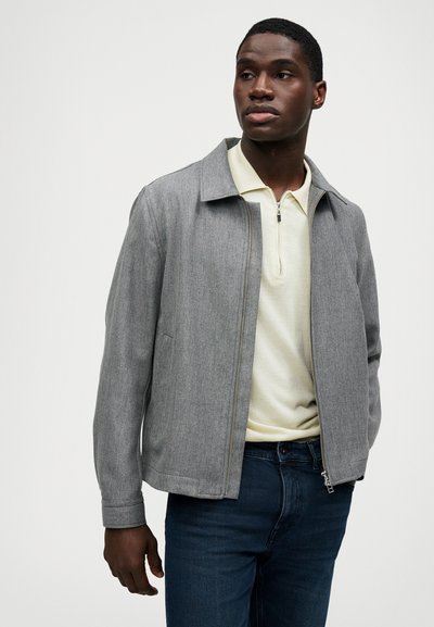 Casaco cinza com fecho éclair em tecido leve, com um colarinho clássico, combinado com uma camisa polo amarelo-claro e jeans azul-escuro.