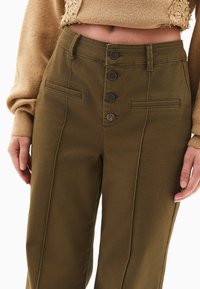 Oliwkowe spodnie z wysokim stanem, z pięcioma guzikami z przodu, dwiema bocznymi kieszeniami i widocznymi detalami ściegów wzdłuż nóg.