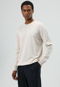 Lichtbeige trui met lange mouwen, ronde hals en een gestructureerde stof; smalle pasvorm met geribbelde manchetten, gecombineerd met een donkere broek.