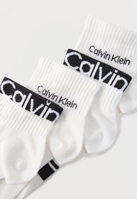 Τρία ζευγάρια λευκές κάλτσες κοντού μήκους με μαύρα λογότυπα Calvin Klein στις ριγωτές μανσέτες, τακτοποιημένα να επικαλύπτονται σε ανοιχτό φόντο.