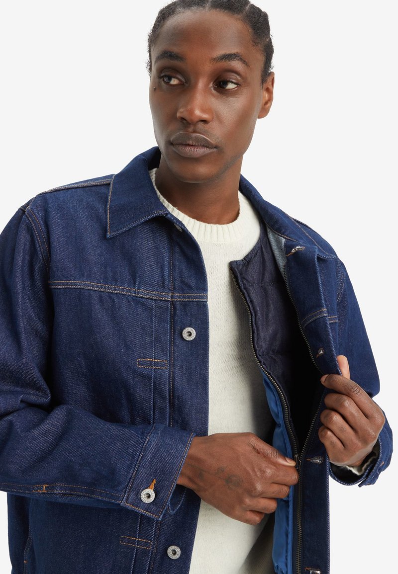 Jeansjacke mit dunkelblauer Oberfläche, traditionellem Kragen, Druckknöpfen und innerem blauen Futter. Verfügt über einen Reißverschluss und sichtbare Nähte.