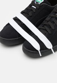 Schwarze Wildledersneaker mit weißen flachen Schnürsenkeln; Gummisohle mit gerippter Textur; Logotag sichtbar auf der Zunge.