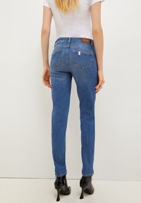 Mujer vestida con jeans ajustados de talle alto, de color azul, con bolsillos traseros y botas de tacón negro hasta el tobillo, de pie contra un fondo claro y liso.