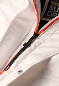 Witte buitenjas met een oranje rits en een zwarte trekker; heeft merkvermelding en een zachte, gladde textuur. De binnenvoering is donker.