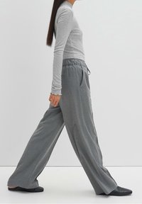 Personne portant un col roulé gris et un pantalon gris ample, marchant en chaussures noires sur un fond clair uni.