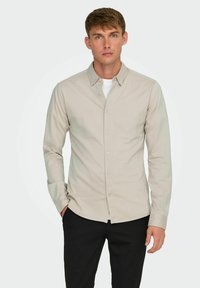 Camisa beige clara de botones con una textura suave, mangas largas, un cuello clásico y botones visibles, combinada con pantalones negros.