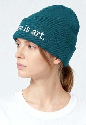 Bonnet tricoté bleu sarcelle avec une texture côtelée, arborant le slogan "she is art." brodé en blanc. Porté par un modèle sur un fond neutre.