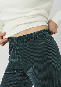 Pantalon en velours côtelé bleu sarcelle avec côtes verticales, taille élastique et un léger effet scintillant, accompagné d'un pull crème au-dessus de la taille.