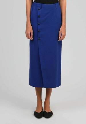 Femme portant une jupe bleue taille haute, longueur genou, avec des boutons noirs sur le côté gauche et des ballerines noires pointues.