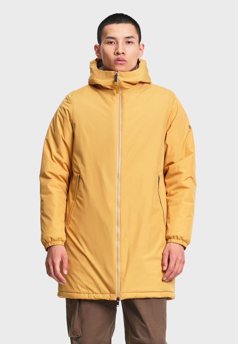Homme portant une longue veste d'hiver à capuche jaune moutarde avec une fermeture éclair à l'avant et des poches latérales, debout devant un fond clair uni.