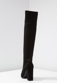 Bota negra a la altura de la rodilla con un upper de punto texturizado y un tacón de ante. Diseño de tacón bloque, que presenta un puño acanalado y un patrón en Chevron en el cañón.