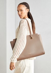 Beige leather tote bag with a structured shape and gold hardware details. Worn with a textured cream sweater, showcasing knitted design.