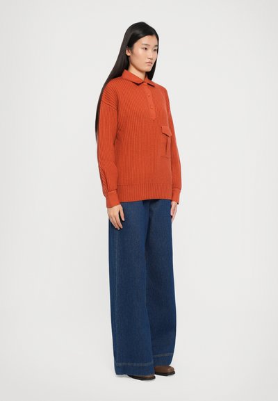 Roestkleurige gebreide trui met een polo kraag, voorzien van een voorzak en een geribbelde textuur, gecombineerd met brede blauwe denim broek.