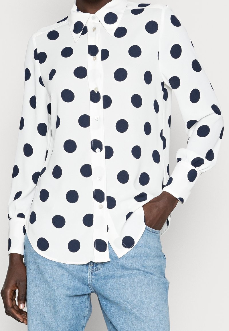 Biała, długa koszula z guzikiem z dużymi czarnymi kropkami, noszona na luzie nad jasnoniebieskimi dżinsami, z jedną ręką w kieszeni.