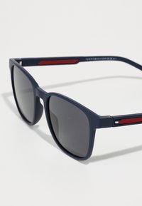 Ochelari de soare navy cu lentile pătrate și nuanță gri închis; prezintă un accent roșu pe brațe, o suprafață texturată și detalii cu logo pe tâmpla.