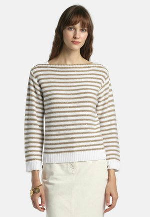 Barna hajú nő fehér és bézs csíkos pulóverben és fehér szoknyában, egyszínű világos háttér előtt állva.