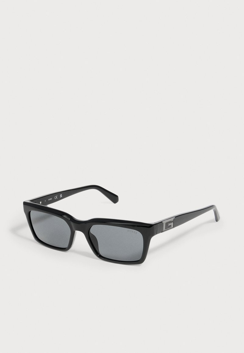 Lunettes de soleil rectangulaires noires avec verres foncés et logo G argenté sur les branches, posées sur une surface claire.