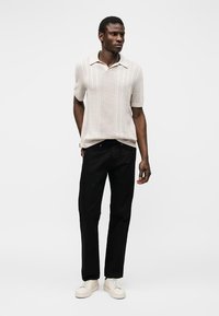 Άνδρας στέκεται φορώντας μπεζ κοντομάνικο πλεκτό πόλο, μαύρο παντελόνι και λευκά αθλητικά παπούτσια σε απλό φωτεινό φόντο.