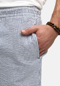Kratke hlače od pamuka s plavo-bijelim prugastim uzorkom, elastičnom strukom i bočnim džepom. Ruka odmara u džepu, vidljivo kontrastno narukvica.