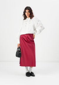 Biela blúzka s vyšívanými detailami, skombinovaná s dlhou burgundskou saténovou sukňou. Čierna kabelka a mokasíny dotvárajú outfit. Viditeľné šedé ponožky.